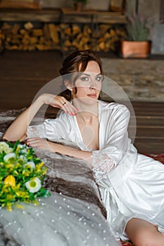 A beautiful young brunette in a silk shirt sits next to the bed with a bouquet of flowers and a wedding veil. Bride`s morning.