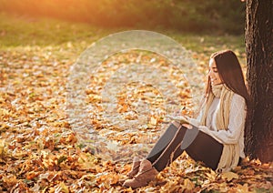 Beautiful young brunette sitting on a fallen autumn leaves in a park, reading a book or write a diary