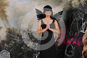 Beautiful young brunette woman in black angel costume with wings over dark spooky background in praying pose. Halloween