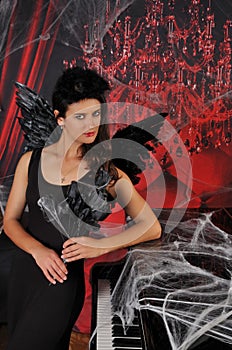 Beautiful young brunette woman in black angel costume with wings over spooky red background posing by piano with rose