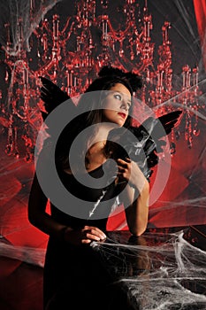 Beautiful young brunette woman in black angel costume with wings over spooky red background posing by piano with rose