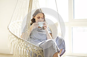 Beautiful young brunette woman drinking coffee while relaxing in knitted macrame style rocking chair