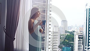 Beautiful young brunette woman drinking coffee stands by the panoramic window with amazing city view. slow motion