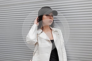 Beautiful young brunette woman in a fashionable white leather jacket in a T-shirt in a stylish black baseball cap posing