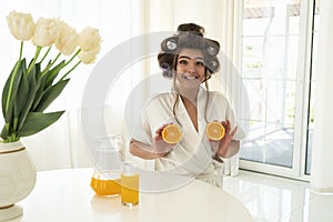 Beautiful young brunette woman in hair curlers holding oranges in both hands near her breasts in bright kitchen looking surprised