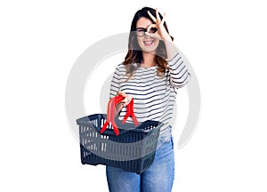 Beautiful young brunette woman holding supermarket shopping basket smiling happy doing ok sign with hand on eye looking through