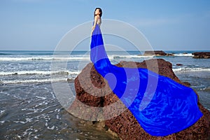 Beautiful and young brunette woman posing in a long dress blue chameleon color train chic perfume on the seashore near