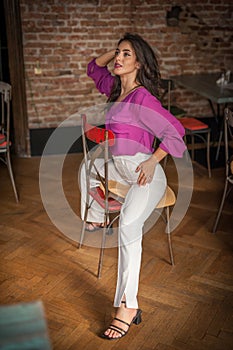 Beautiful young brunette woman in a purple blouse and white pants posing indoor