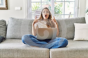 Beautiful young brunette woman sitting on the sofa using computer laptop at home amazed and surprised looking up and pointing with