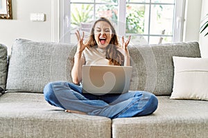 Beautiful young brunette woman sitting on the sofa using computer laptop at home celebrating mad and crazy for success with arms