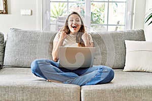 Beautiful young brunette woman sitting on the sofa using computer laptop at home celebrating surprised and amazed for success with