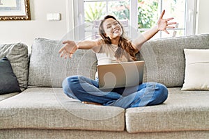 Beautiful young brunette woman sitting on the sofa using computer laptop at home looking at the camera smiling with open arms for