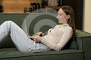 Beautiful young brunette woman using a laptop to work in the living room lying on a green sofa