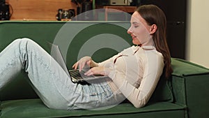 Beautiful young brunette woman using a laptop to work in the living room lying on a green sofa