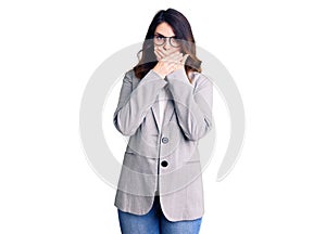 Beautiful young brunette woman wearing business clothes and glasses shocked covering mouth with hands for mistake