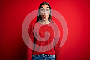 Beautiful young brunette woman wearing casual sweater standing over red isolated background afraid and shocked with surprise