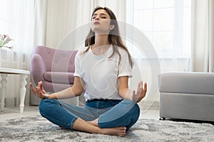 Beautiful young brunette woman wearing jeans and white t-shirt sitting on the floor in asana yoga position in bright livingroom