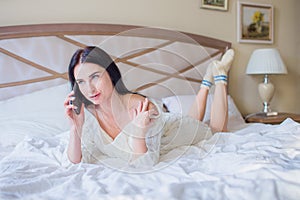 Beautiful young brunette woman in white knitted dress talking on the phone while lying in bed at home or in hotel and laughing