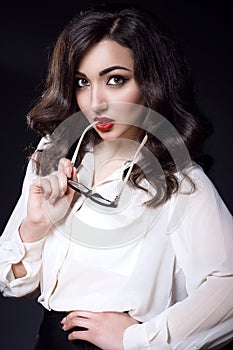 Beautiful young business woman with dark wavy hair and red lips wearing white silk blouse keeping the side of glasses in her mouth