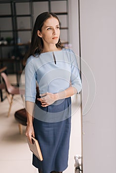 Beautiful young business woman holding file folder while standing near window in modern office room.