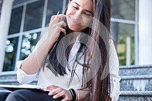 Beautiful young business woman of indian nationality, straightens her hair, sits on the steps, smiles, and writes in pencil in a