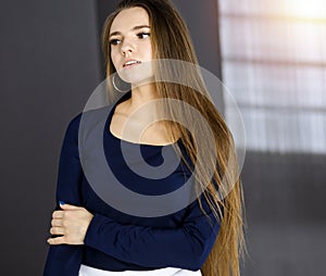 Beautiful young business woman with long hair or female student urban dressed is standing straight and posing at camera