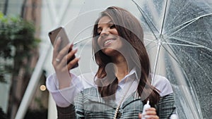 Beautiful young business woman using smartphone on the street in rainy weather, smiling, holding umbrella, communication