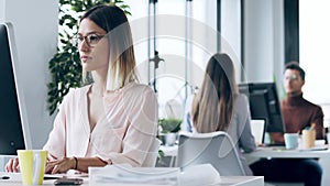 Beautiful young business woman working with computer while cheeking some documents sitting in desk in modern startup off