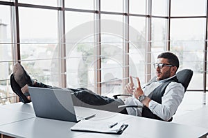 Beautiful young businessman with glasses holding his legs on the table looking at a laptop in the office