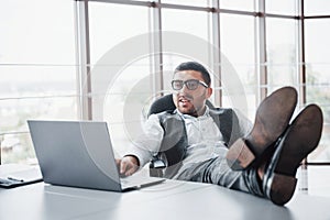 Beautiful young businessman with glasses holding his legs on the table looking at a laptop in the office