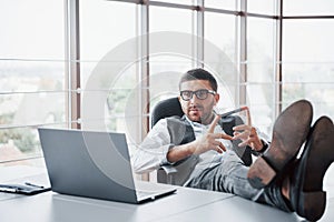 Beautiful young businessman with glasses holding his legs on the table looking at a laptop in the office