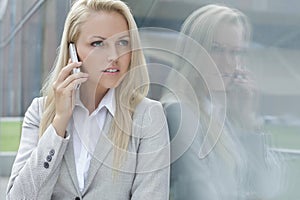 Beautiful young businesswoman conversing on cell phone while looking away by glass wall