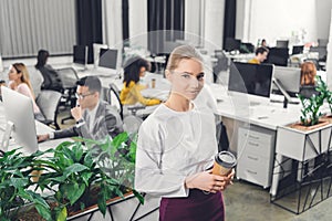Beautiful young businesswoman holding coffee to go and smiling at camera while colleagues working behind