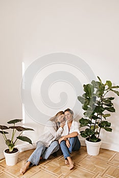 Beautiful young caucasian blonde leans on her mother's shoulder sitting on floor in spacious room.