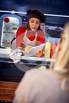 Beautiful young caucasian female employee in fast food service taking order from a customer, writing down on a paper