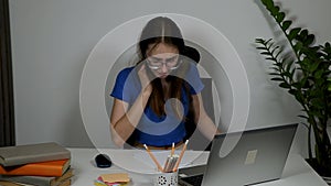 a beautiful young Caucasian girl in glasses looks carefully at the laptop screen while sitting at a white table. online