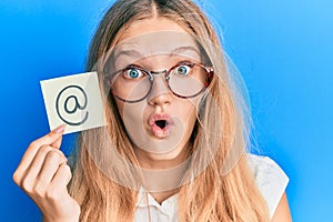 Beautiful young caucasian girl holding online mail symbol on paper scared and amazed with open mouth for surprise, disbelief face