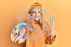 Beautiful young caucasian girl holding paintbrush and painter palette smiling and laughing hard out loud because funny crazy joke