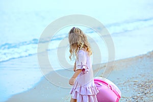 Beautiful young Caucasian girl on the sea blue coast in a dress light pale pink with a children`s pink suitcase for holidays