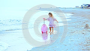 Beautiful young Caucasian girl on the sea blue coast in a dress light pale pink with a children`s pink suitcase for holidays