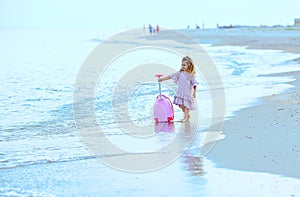 Beautiful young Caucasian girl on the sea blue coast in a dress light pale pink with a children`s pink suitcase for holidays