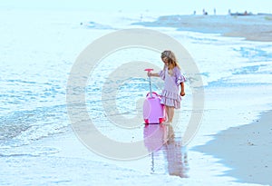 Beautiful young Caucasian girl on the sea blue coast in a dress light pale pink with a children`s pink suitcase for holidays