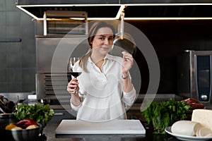 Beautiful young caucasian girl standing in kitchen in a white uniform smiling and tasting red wine