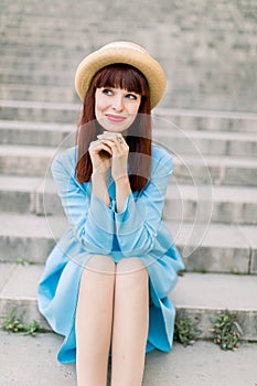 Beautiful young caucasian girl in urban background, sitting on stairs. Pretty red haired girl wearing hat and blue dress