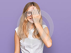 Beautiful young caucasian girl wearing casual clothes and glasses covering one eye with hand, confident smile on face and surprise