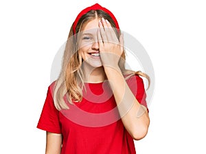 Beautiful young caucasian girl wearing casual red t shirt covering one eye with hand, confident smile on face and surprise emotion