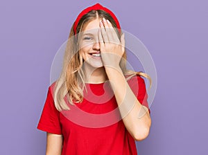 Beautiful young caucasian girl wearing casual red t shirt covering one eye with hand, confident smile on face and surprise emotion