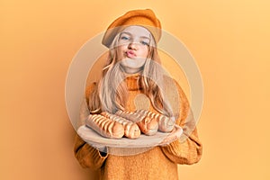 Beautiful young caucasian girl wearing french look with beret holding baguettes bread looking at the camera blowing a kiss being