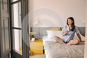 Beautiful young caucasian girl wears pajamas looking out window sits on bed.