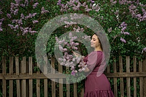 A beautiful young Caucasian smiling happy girl in purple dress near a Bush of blooming lilac. Model plus size. Spring